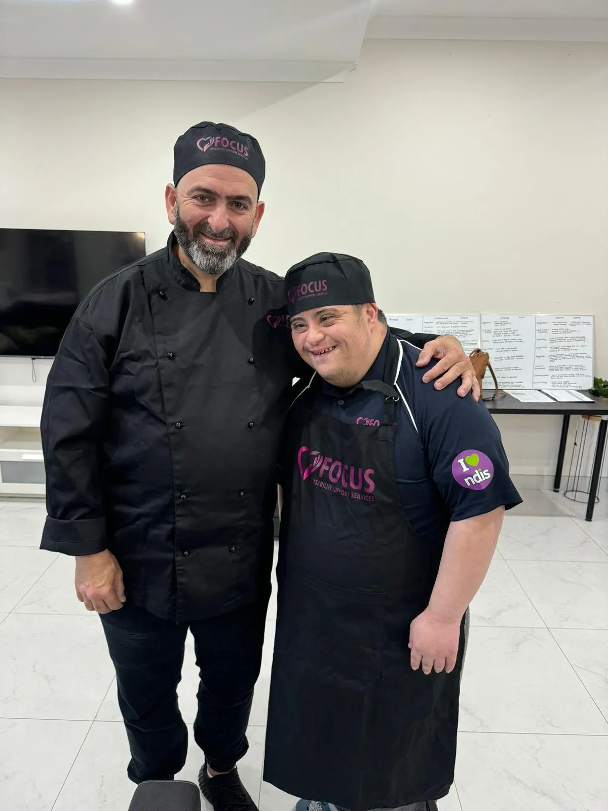 A Focus support worker in chef's whites with his arm around a participant in a Focus apron, in the group centre kitchen.