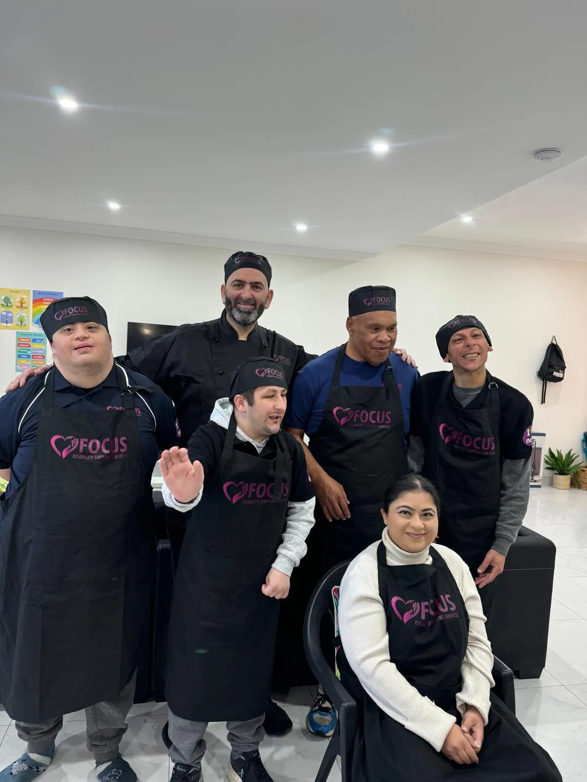 Seven members of the Focus crew — participants and support workers in Focus aprons and caps — standing together in the group centre kitchen.